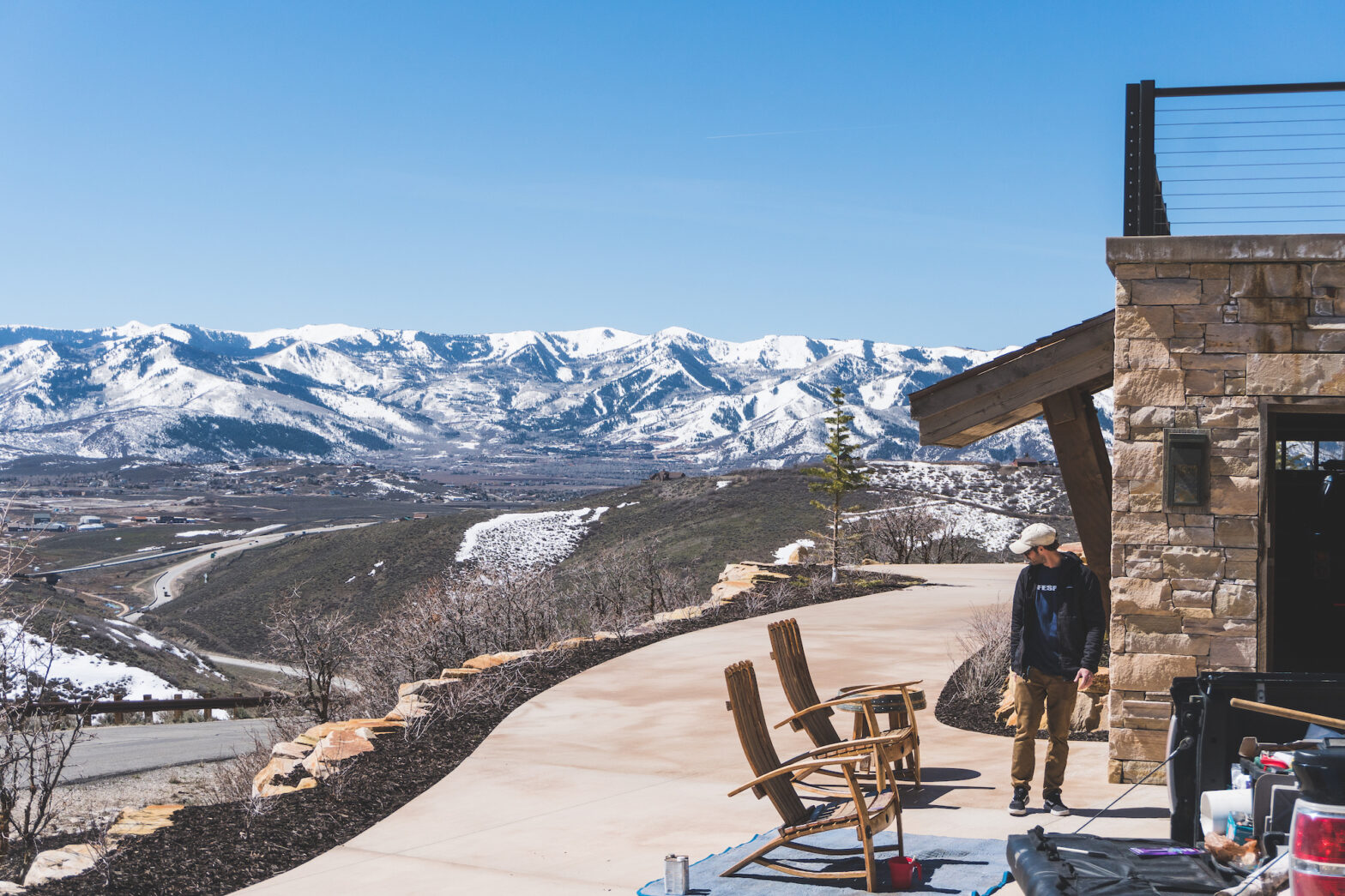 Spring Maintenance - A man in a hat, walking by chairs on a driveway, towards tools. Snow-capped mountains in the background.