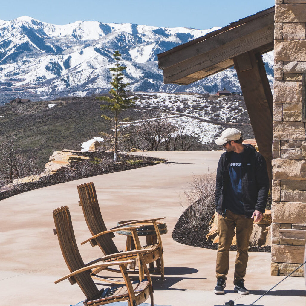 A man in a hat, walking by chairs on a driveway, towards tools. Snow-capped mountains in the background.
